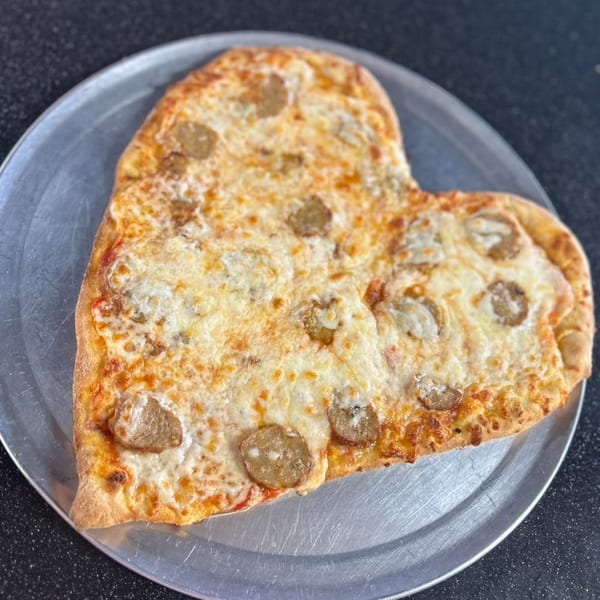 Heart-shaped pizza with melted cheese and sausage slices on a metal pizza pan.
