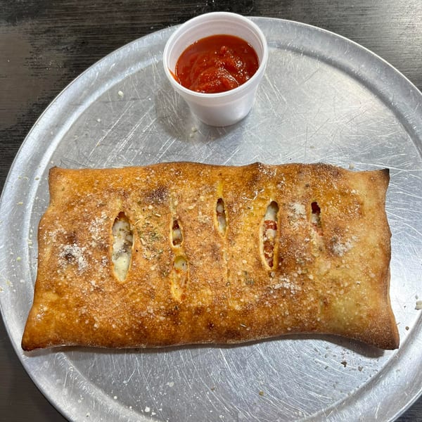 A golden-brown calzone with visible fillings and a side of marinara sauce on a metal tray.